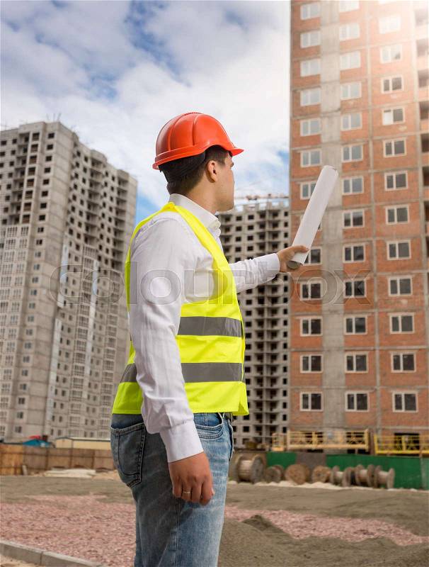 Architect in hardhat pointing with rolled blueprints at building under construction, stock photo