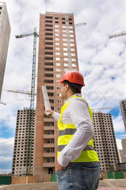Rear view of architect pointing with blueprints at building under construction, stock photo