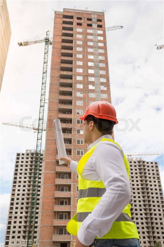 Young architect in hardhat pointing at building under construction, stock photo