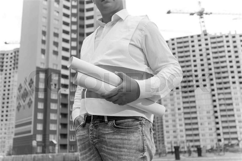 Closeup black and white photo of engineer holding rolled blueprints in background of building site, stock photo