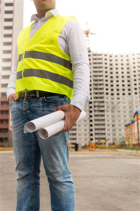 Construction engineer standing at building site and holding blueprints, stock photo