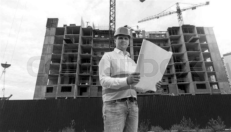 Black and white portrait of young engineer in hardhat with blueprints on building site, stock photo