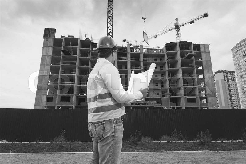 Black and white image of engineer in hardhat and vest inspecting building site, stock photo