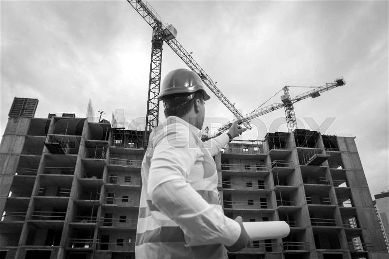 Black and white image of young engineer holding blueprints and pointing at building site with hardhat, stock photo