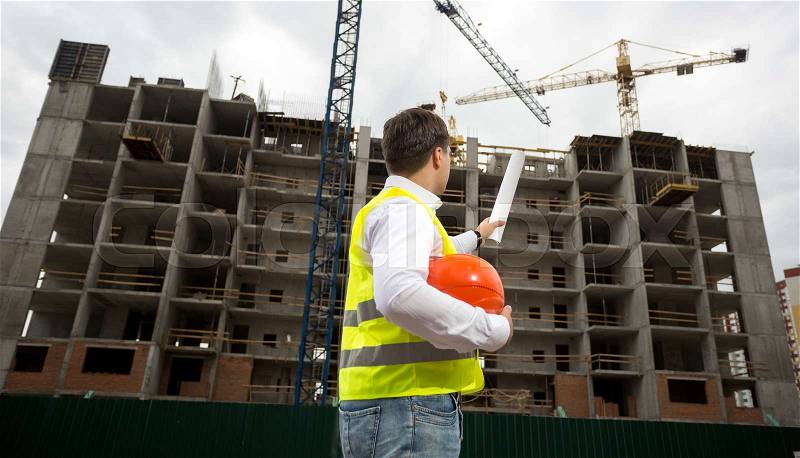 Young engineer pointing at building with rolled blueprints, stock photo