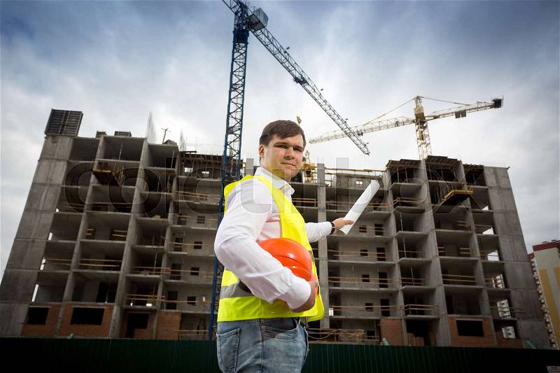 Young engineer in safety vest with blueprints at building site, stock photo