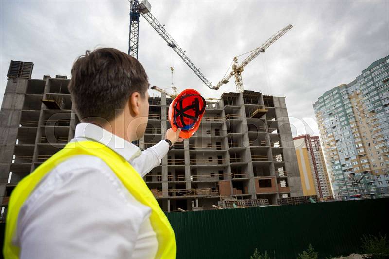Young construction engineer pointing at building under construction with red hardhat, stock photo