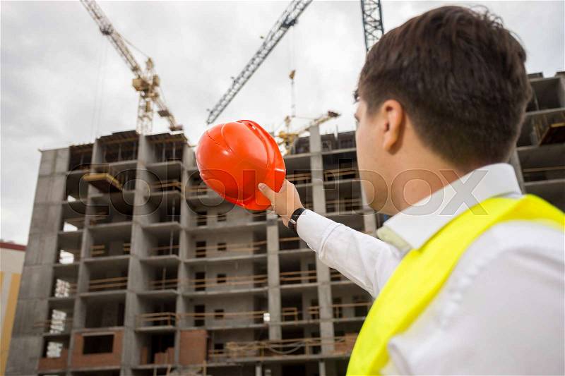 Young construction engineer pointing at building under construction with red hardhat, stock photo