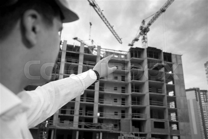 Closeup black and white image of construction engineer pointing with hand at building under construction, stock photo