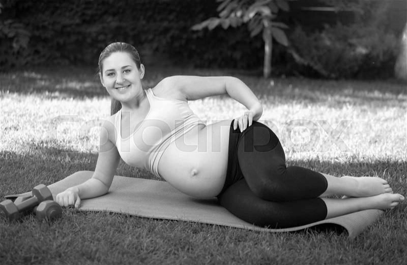 Black and white image of young expectant mother stretching before yoga on grass at park, stock photo