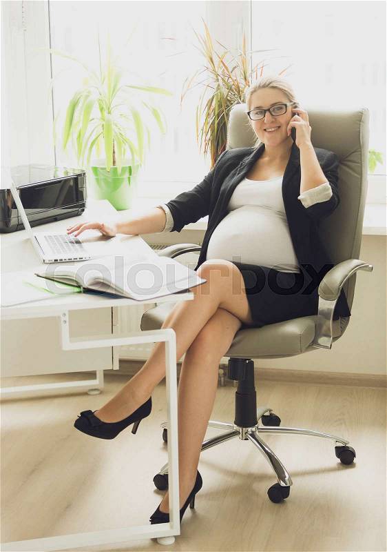 Beautiful smiling pregnant woman in black suit talking by phone at office, stock photo