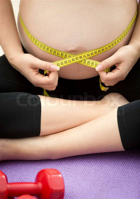Closeup photo of pregnant woman sitting on fitness mat and measuring belly with tape, stock photo