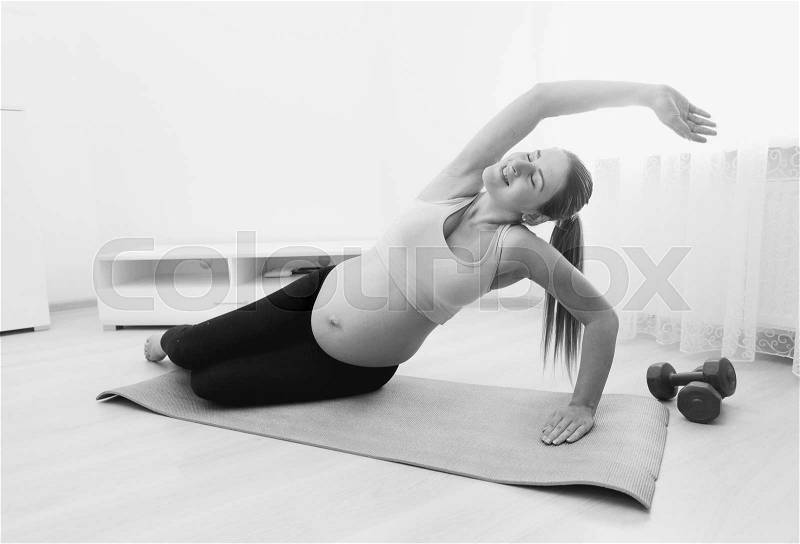 Black and white image of beautiful pregnant woman stretching on fitness mat at home, stock photo
