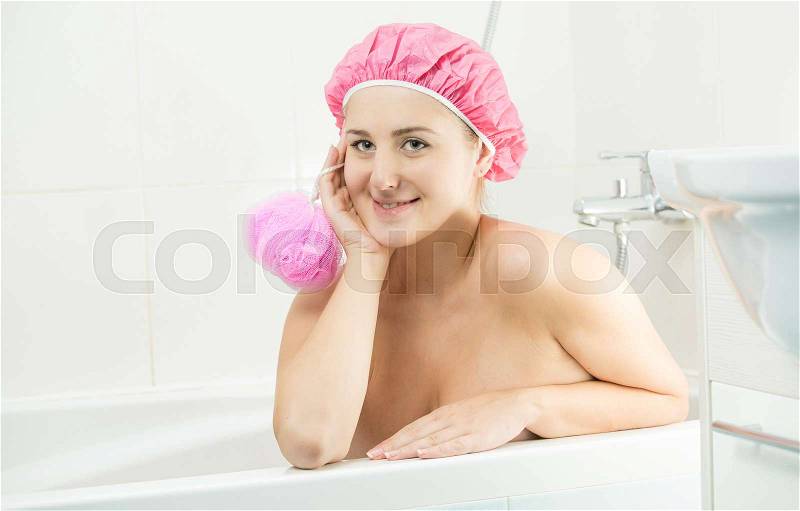 Beautiful smiling woman wearing shower cap lying in bath and looking at camera, stock photo