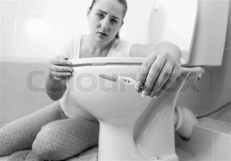 Black and white portrait of sad sick woman sitting in bathroom and holding positive pregnancy test, stock photo