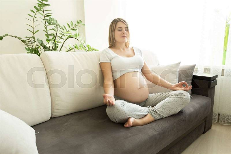 Young pregnant woman practicing yoga on sofa at living room, stock photo