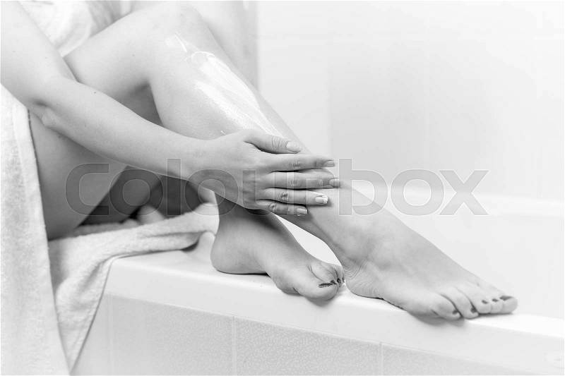 Black and white image of young woman applying moisturizing creme on legs after bathing, stock photo