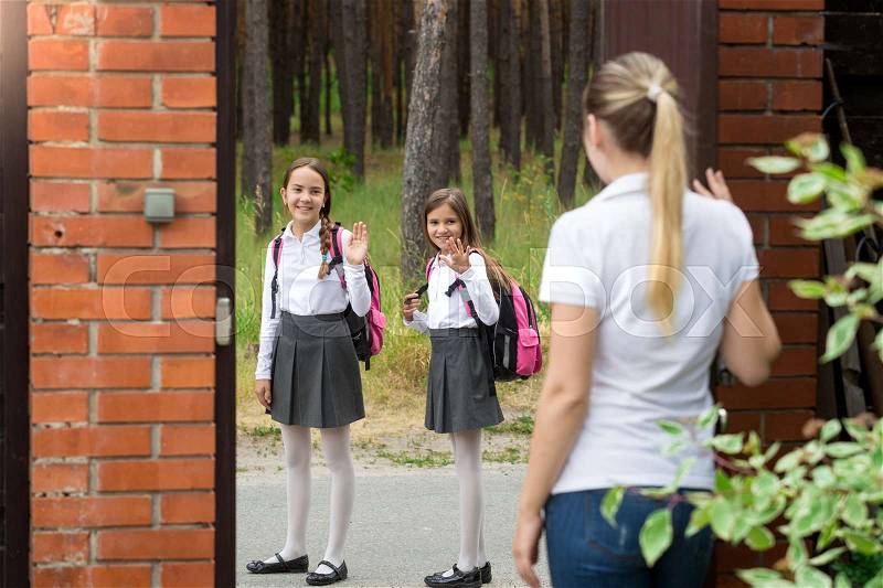 Mother standing in house yard and waving to her daughters walking to school, stock photo