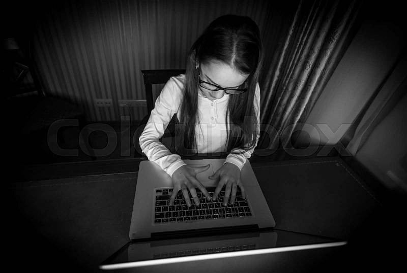 Black and white portrait of teenage girl sitting in dark room with laptop, stock photo