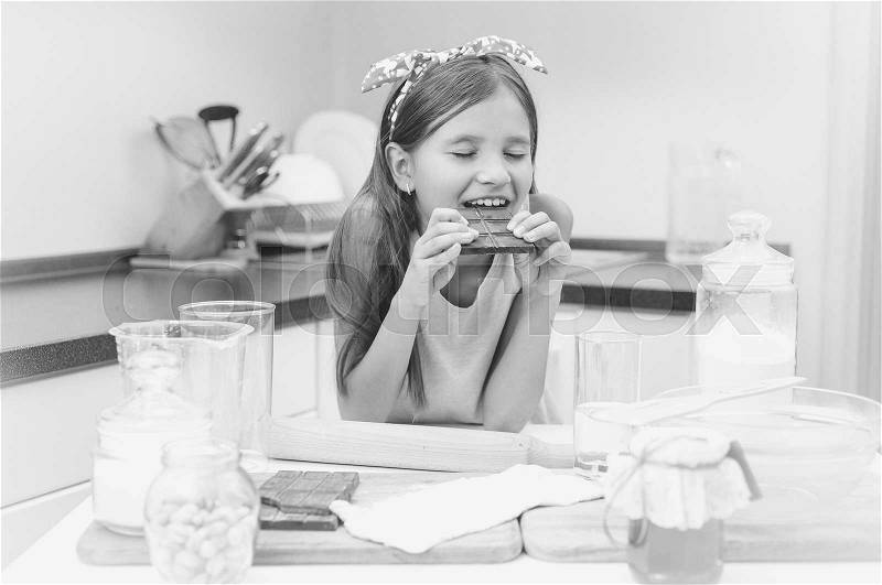 Black and white portrait of cute girl leaning on kitchen table and eating chocolate, stock photo