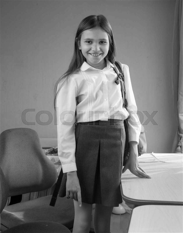 Black and white portrait of cute girl with school bag standing behind desk at bedroom, stock photo