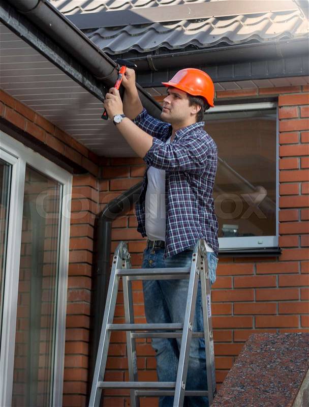 Worker standing on step ladder and repairing gutter on house, stock photo