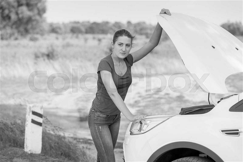 Black and white image of young woman opening hood of the broken car at roadside, stock photo