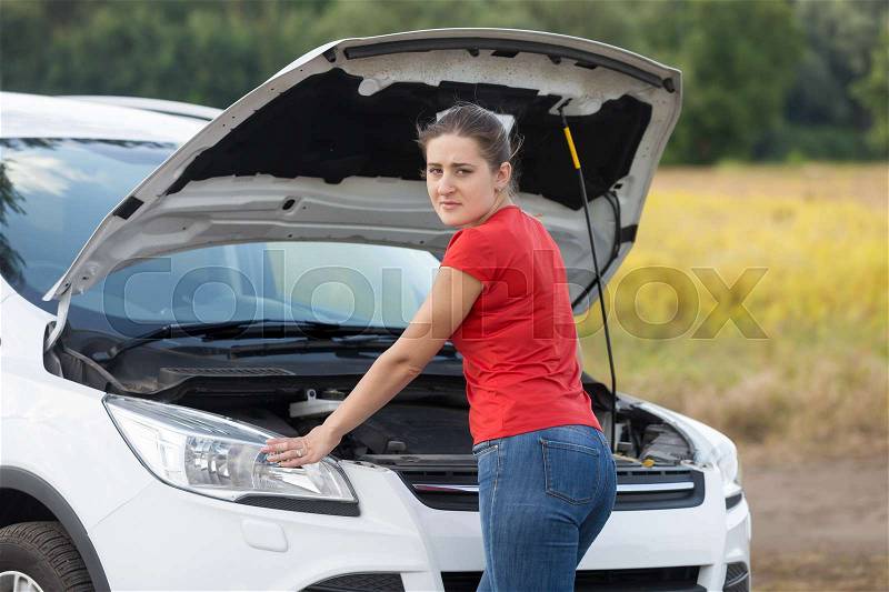 Young woman looking under the hood of broken car at rural road, stock photo