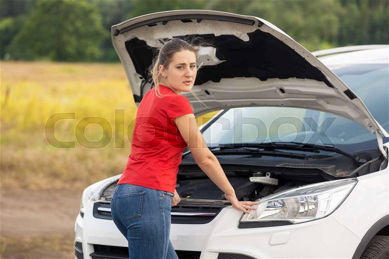 Portrait of young woman looking under the hood of broken car, stock photo