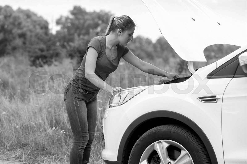 Black and white image of young woman looking under the hood of overheated car at meadow, stock photo