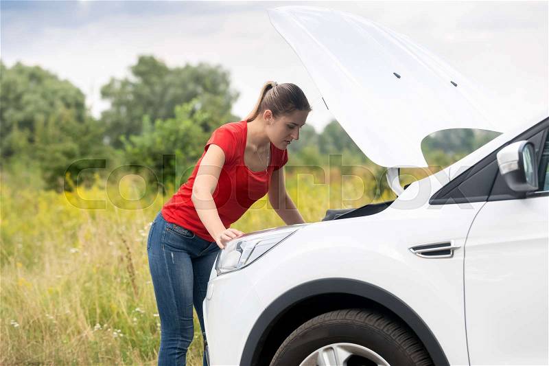 Woman looking under the hood of overheated car at meadow, stock photo