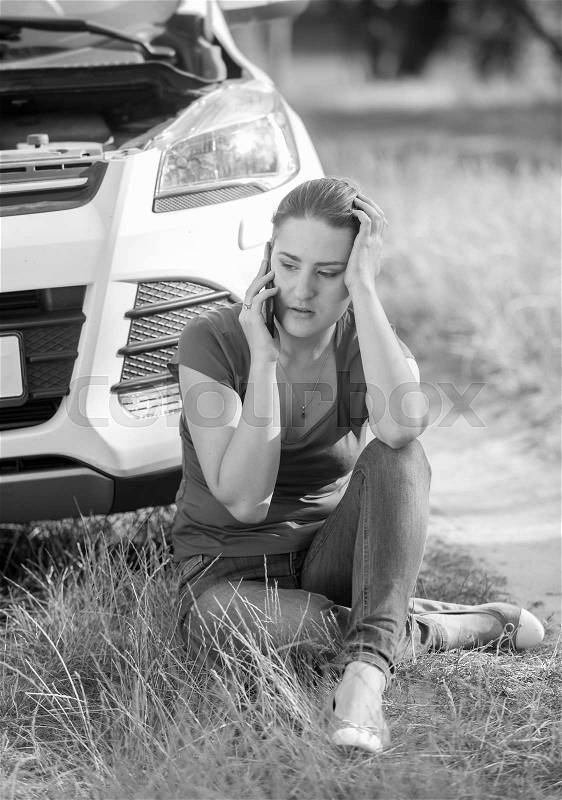 Black and white image of young woman sitting on ground and leaning on broken car, stock photo