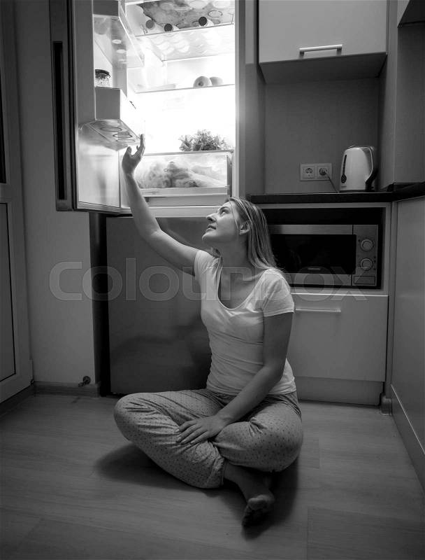 Black and white image of young woman sitting on floor and reaching for food from open refrigerator at late night, stock photo
