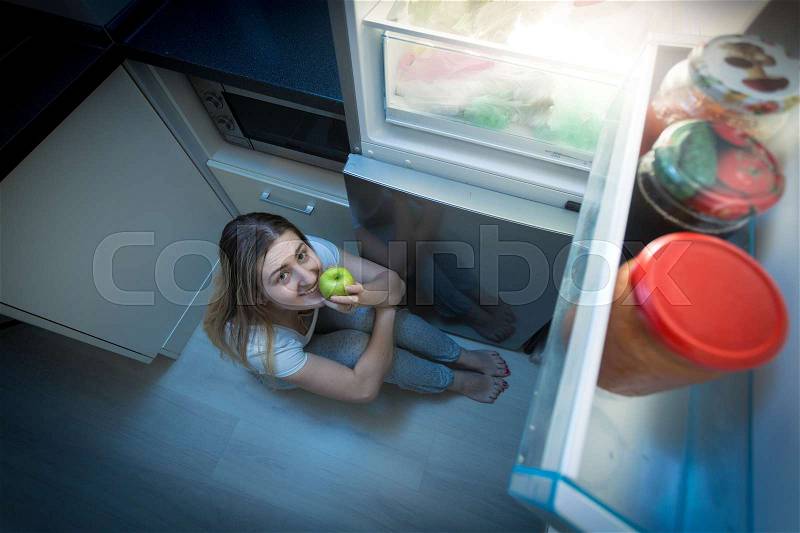 Hungry woman eating apple on kitchen floor at late night, stock photo
