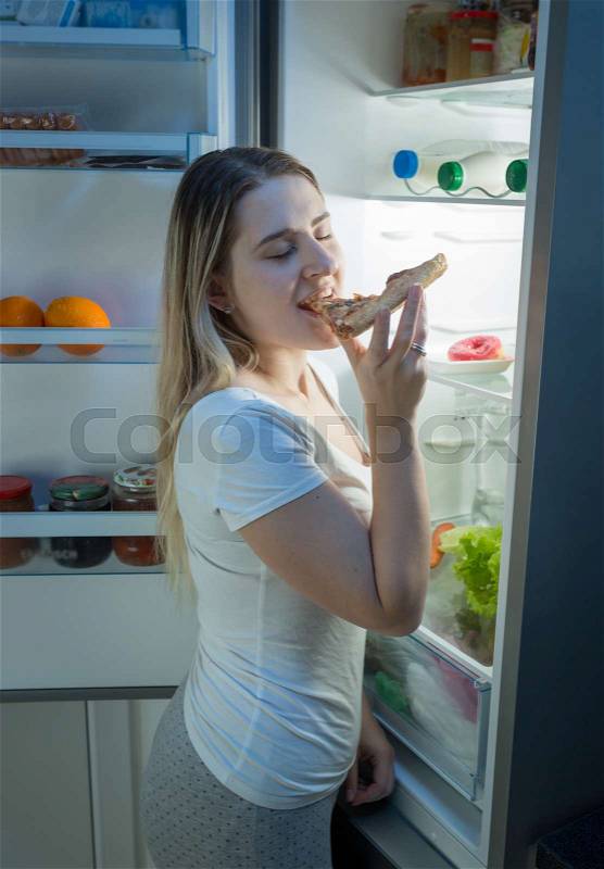 Young smiling woman eating pizza on kitchen at night, stock photo