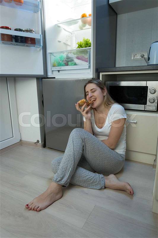 Young sleepless woman sitting on kitchen floor and eating pizza, stock photo