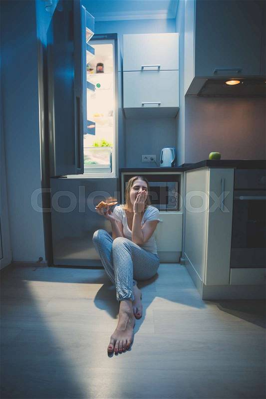 Sleepy woman sitting on floor at open fridge at late night, stock photo