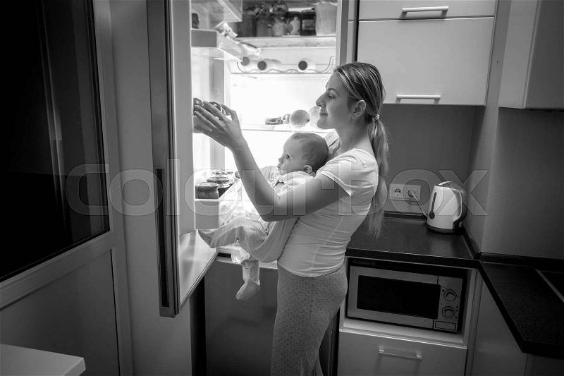 Black and white image of young mother and baby son looking inside the fridge for something to eat, stock photo