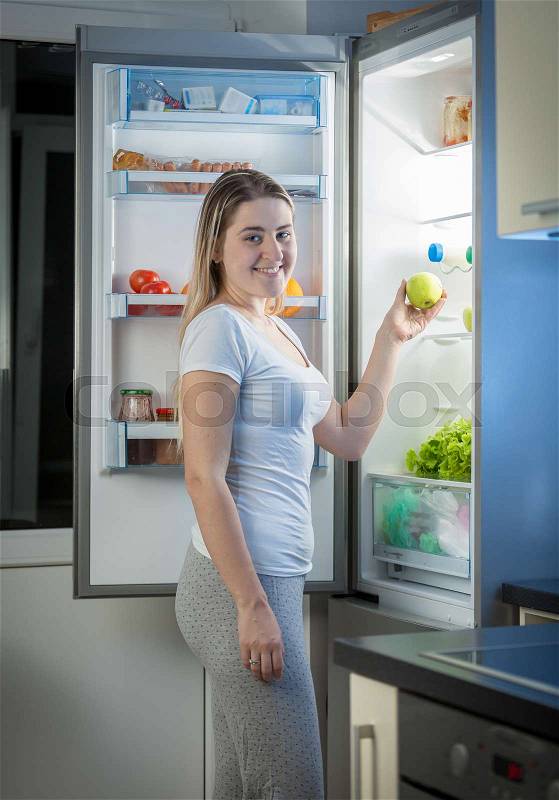 Smiling woman taking green apple from fridge at night, stock photo