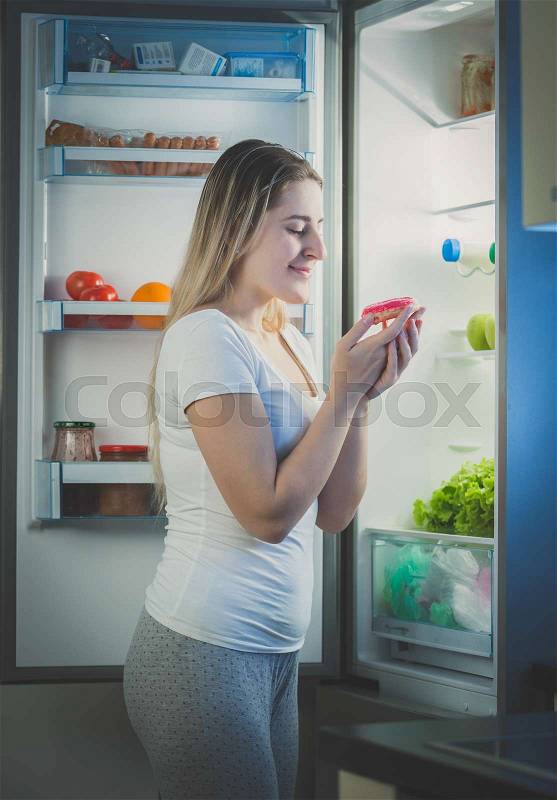 Beautiful woman taking donut from fridge at late evening, stock photo