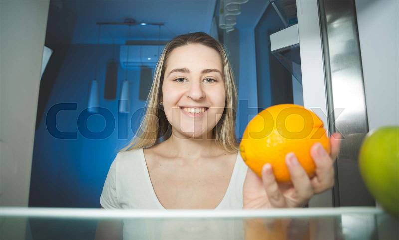 Beautiful smiling woman holding orange and looking inside the refrigerator, stock photo