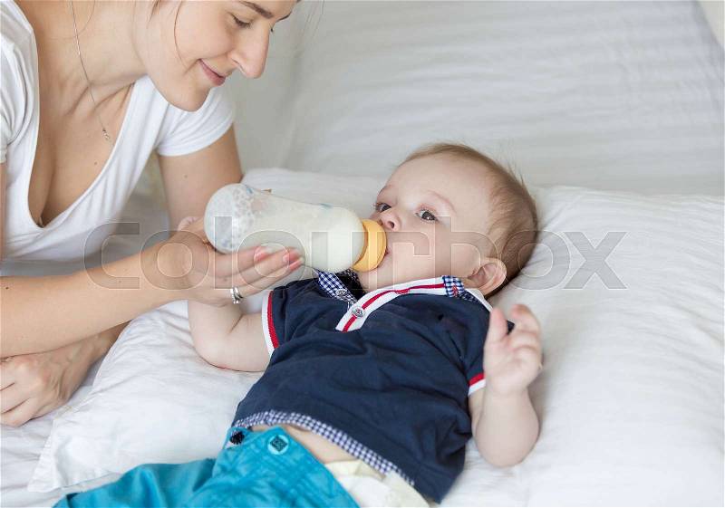 Young woman feeding her baby son from milk bottle on bed, stock photo
