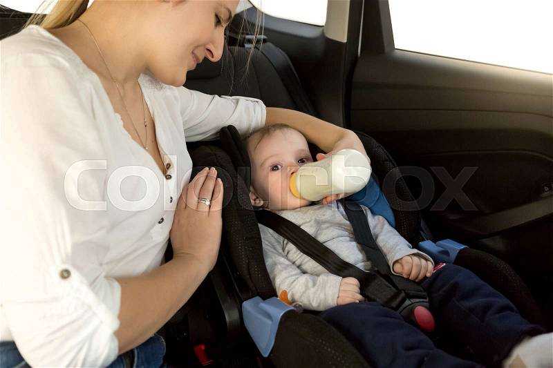 Mother feeding baby boy with milk on car back seat, stock photo