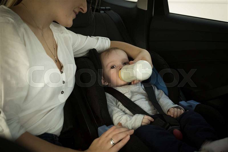 Young mother giving milk in bottle to her little son in car, stock photo