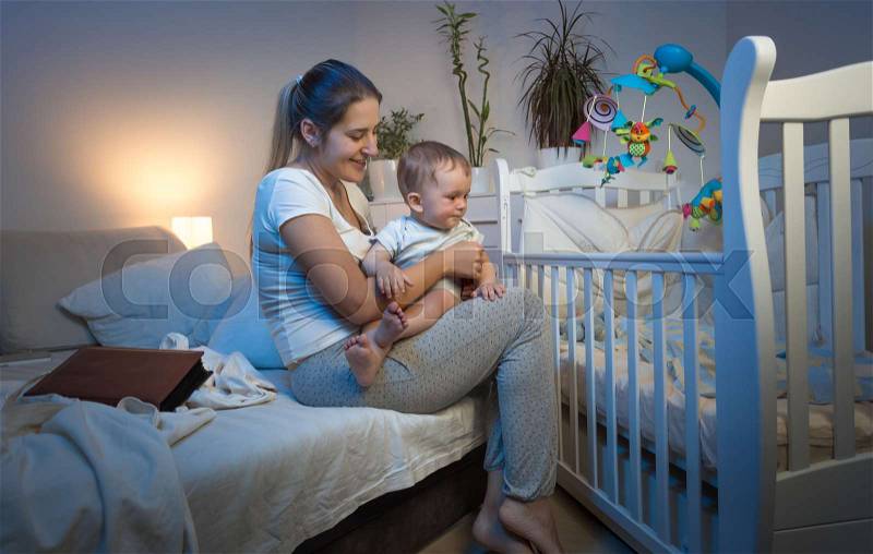 Beautiful young mother taking baby out of crib at night, stock photo
