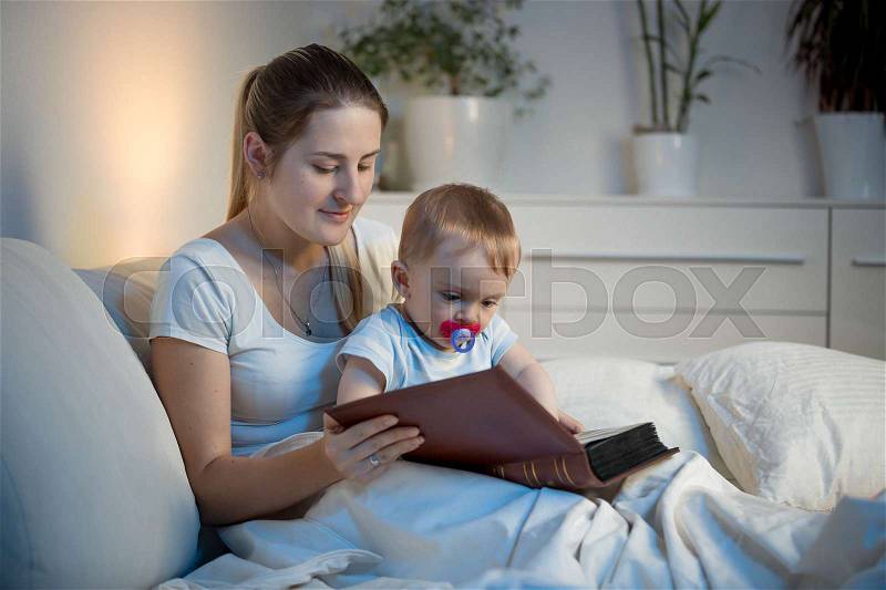 Beautiful smiling mother telling story to her baby at bed before going to sleep, stock photo