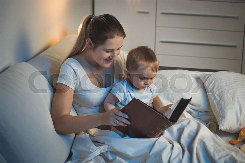 Young mother storytelling to her adorable baby boy at bed, stock photo