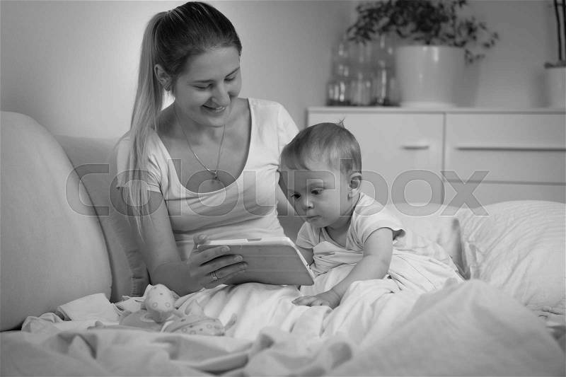 Black and white image of baby with mother using tablet computer in bed at night, stock photo