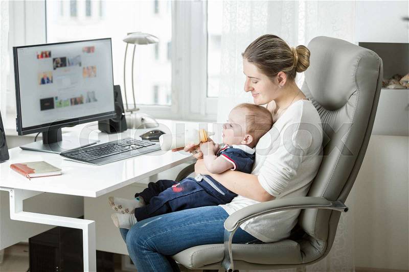 Smiling mother working at home office and feeding her baby from bottle, stock photo