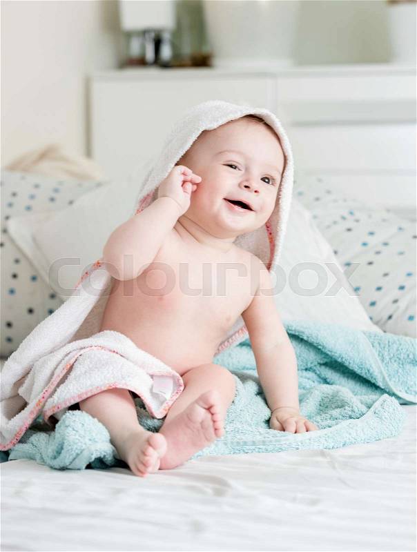 9 months old baby boy sitting on bed covered in towel after bathing, stock photo